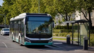 Modern lackierter Stadtbus in Schwarz und Türkis an einer Bushaltestelle mit Glasüberdachung auf einer Straße mit Bäumen