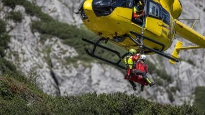 Gelber Rettungshubschrauber transportiert verletzte Person in roter Trage über Berglandschaft