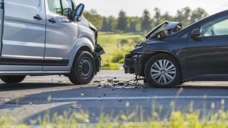 Silberner Kleintransporter und dunkler Pkw nach Frontalkollision auf einer ländlichen Straße.