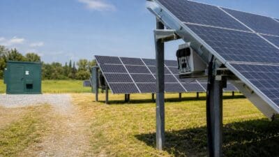 Aufgeständerte Solarpaneele in einer Reihe auf einem grünen Feld neben einem grünen Verteilerkasten unter blauem Himmel.