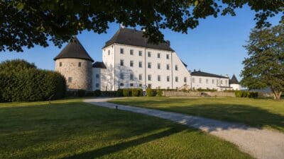 Weißes Schloss mit rundem Steinturm und dunklem Dach hinter Rasen und Kiesweg