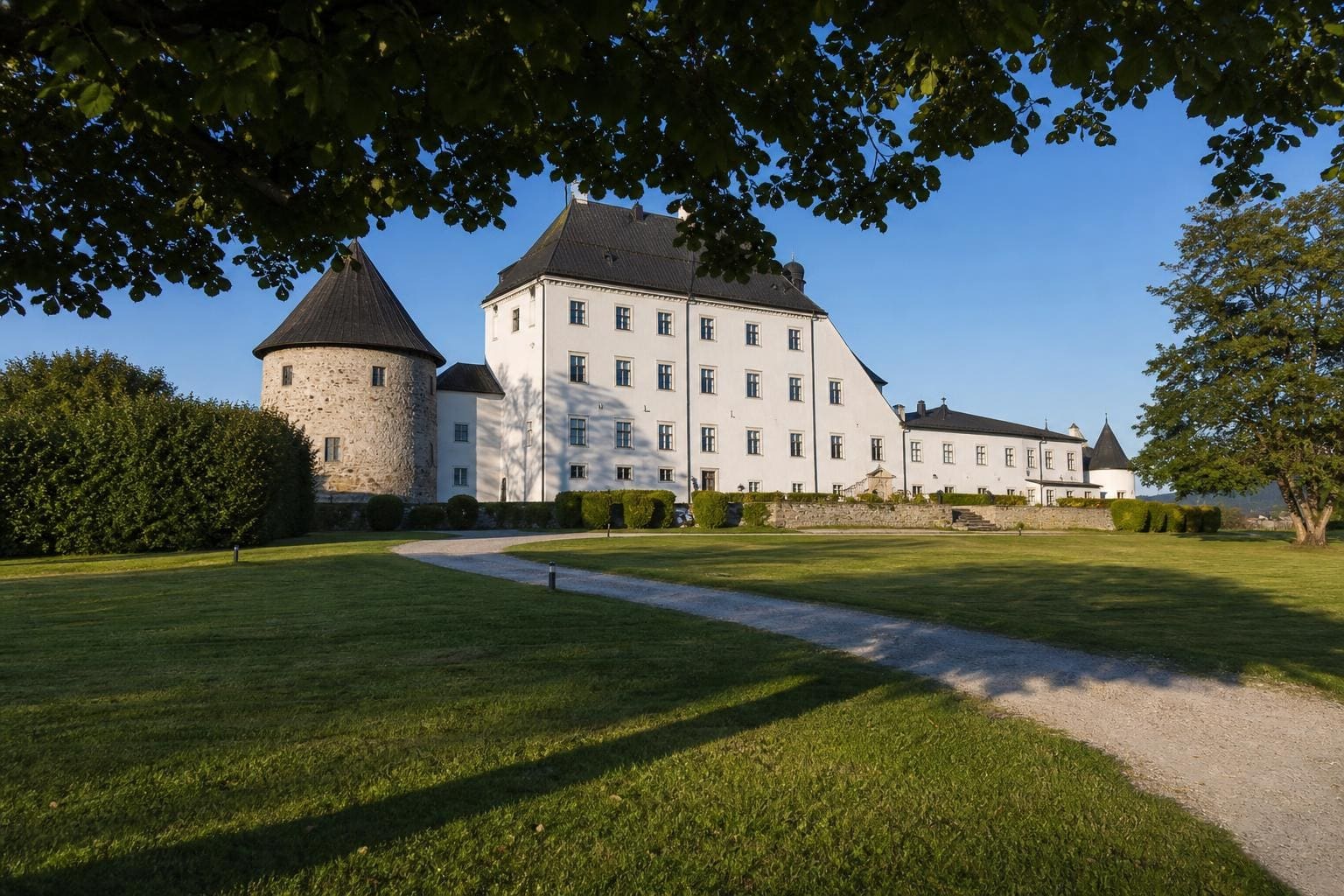 Weißes Schloss mit rundem Steinturm und dunklem Dach hinter Rasen und Kiesweg