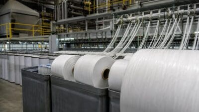 Mehrere große weiße Rollen aus Fasern in grauen Behältern vor einer Produktionsmaschine in einer Fabrikhalle