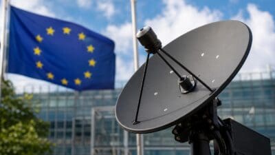 Schwarze Satellitenschüssel im Vordergrund vor einer wehenden EU-Flagge und einem modernen Gebäude