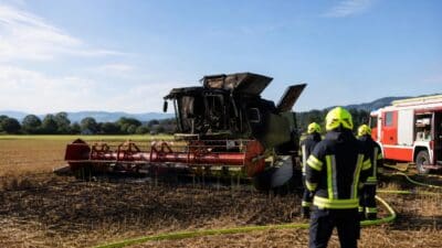 Ausgebrannter Mähdrescher auf einem Feld, davor Feuerwehrleute mit Schläuchen und Löschfahrzeug