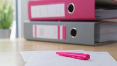 Pinkfarbener Aktenordner auf einem grauen Ordner mit pinkem Stift auf weißem Papier auf einem Holztisch