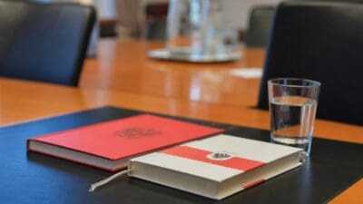 Zwei Bücher mit rotem und weiß-rotem Einband sowie ein Wasserglas auf einem dunklen Tisch.