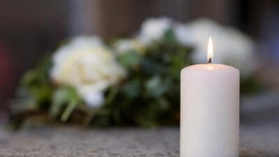 Eine brennende weiße Kerze steht auf einer Steinfläche vor unscharfen weißen Blumen.