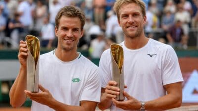 Zwei Tennisspieler in weißen Trikots halten lächelnd goldene Pokale vor Zuschauern.