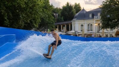 Junger Mann balanciert auf Surfbrett auf spritzender blauer Welle einer FlowRider-Anlage