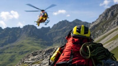 Rettungshubschrauber fliegt über Bergkette mit rotem Rucksack Helm und Seil im Vordergrund