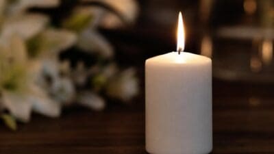 Brennende weiße Stumpenkerze auf dunklem Holz vor unscharfen weißen Blumen