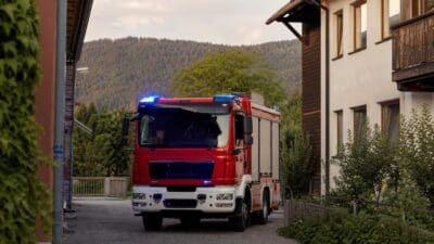 Rotes Feuerwehrfahrzeug mit blauem Blaulicht auf einer Straße in einem Wohngebiet mit Bergen im Hintergrund