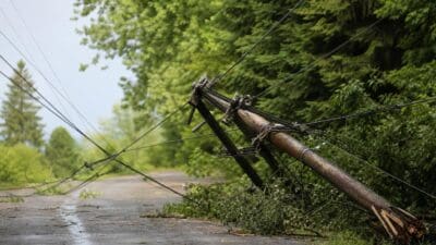 Umgestürzter Holzmast mit herabhängenden Stromleitungen liegt auf nasser Straße vor Bäumen