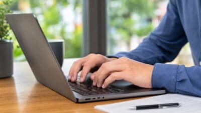 Hände tippen auf der Tastatur eines Laptops auf einem Holztisch mit Dokumenten daneben.