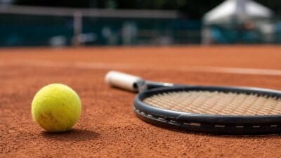 Nahaufnahme eines gelben Tennisballs neben einem Tennisschläger auf rotem Sandplatz