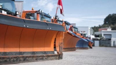 Mehrere orangene Schneeräumfahrzeuge mit rot-weißer Flagge auf einem Platz