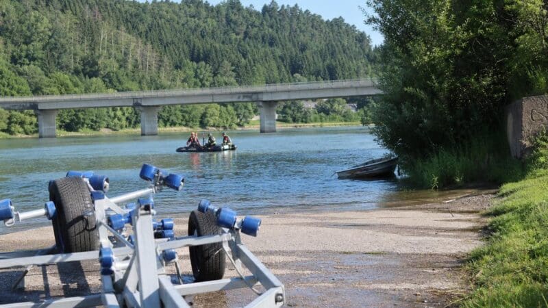 Einsatzkräfte in einem Schlauchboot auf der Drau bei einer Brücke und einem Bootstrailer am Ufer