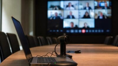 Laptop und Webcam auf einem Holztisch vor einem Bildschirm mit Videokonferenzteilnehmern