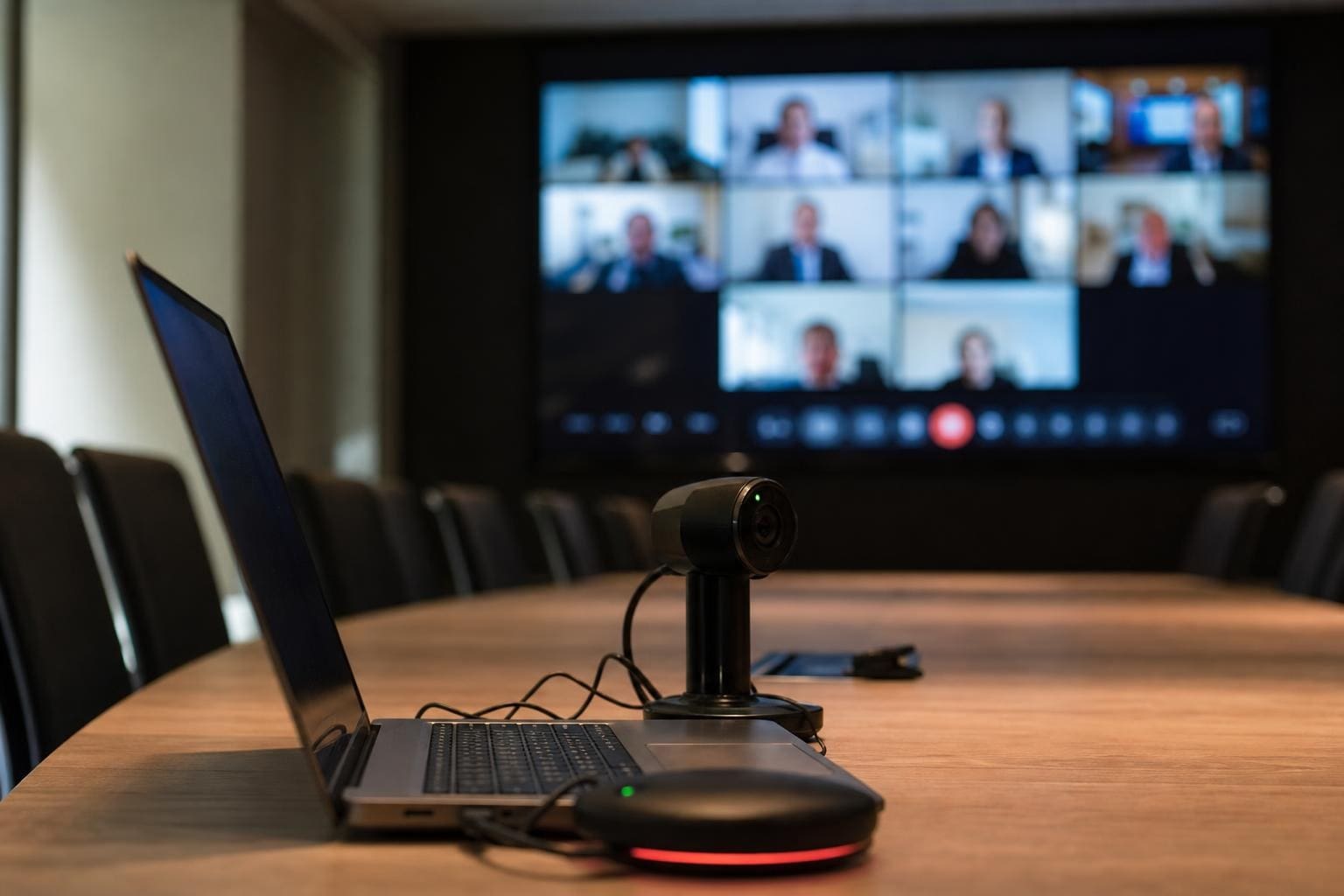 Laptop und Webcam auf einem Holztisch vor einem Bildschirm mit Videokonferenzteilnehmern