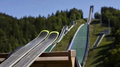 Zwei Skier mit gelben Kanten auf einem Holzgeländer vor Skisprungschanzen unter blauem Himmel