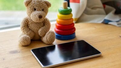Brauner Teddybär sitzt neben buntem Stapelring-Spielzeug und Tablet auf Holztisch