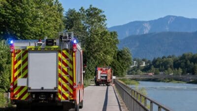 Zwei rote Feuerwehrfahrzeuge mit Blaulicht auf einem Weg am Inn in Schwaz vor Bergen.