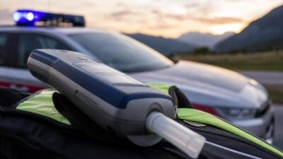 Alkoholtestgerät liegt auf Tasche vor Polizeiauto mit Blaulicht bei Dämmerung
