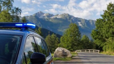 Ein silbernes Einsatzfahrzeug mit aktiviertem blauen Blaulicht steht auf einer Straße vor Bergen.