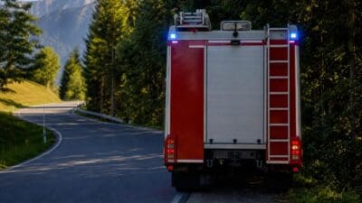 Heckansicht eines roten Feuerwehrfahrzeugs mit blauen Blinklichtern auf einer kurvigen Landstraße in den Bergen