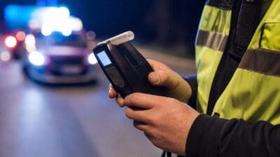 Einsatzkraft in gelber Warnweste bedient ein Alkoholmessgerät vor einem unscharfen Polizeiwagen bei Nacht