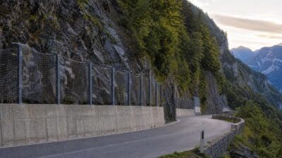 Kurvenreiche Straße am Plöckenpass mit Betonbarriere und Drahtschutznetzen an der Felswand.