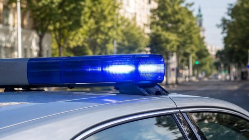 Blaues Blaulicht leuchtet auf dem Dach eines silbernen Polizeiautos vor unscharfer Stadtkulisse.