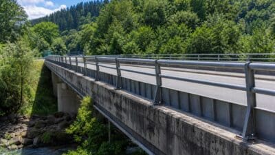 Betonbrücke mit Metallgeländern über einem Fluss vor dicht bewaldeten Hügeln