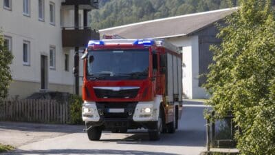 Rotes Feuerwehrfahrzeug mit Blaulicht fährt auf einer Straße in ländlicher Umgebung.