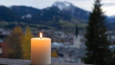 Eine brennende weiße Kerze steht auf einem Holzgeländer vor einer unscharfen Berglandschaft mit Kirchturm