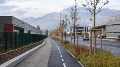 Schwarzer Radweg mit weißen Markierungen neben einer Straße in einem Gewerbegebiet vor Bergkulisse