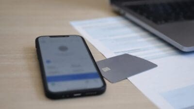 Ein Smartphone und eine Kreditkarte liegen auf Papieren neben einem Laptop auf einem Holztisch.
