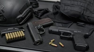 Auf einer dunklen Fläche liegen ein schwarzer Helm, zwei Pistolen, Munition und eine taktische Weste.