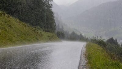 Nasse Bergstraße bei starkem Regen, umgeben von grünen Hängen und nebligen Bergen