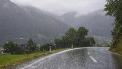 Nasse Landstraße im Starkregen vor bewaldeten Bergen und Häusern im Tal