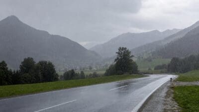 Nasse Landstraße bei starkem Regen vor bewaldeten Bergen und Wiesen
