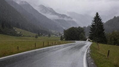 Nasse Landstraße schlängelt sich durch regnerisches Alpen-Tal mit Wiesen und Häusern