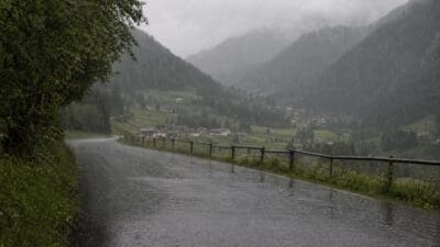 Nasse Landstraße im Regen mit Holzzaun und bewaldeten Bergen im Hintergrund