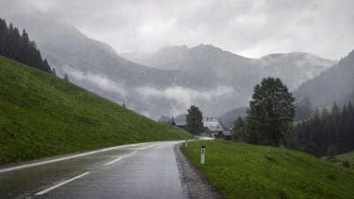 Nasse Landstraße führt bei starkem Regen durch grüne Berglandschaft mit Häusern und Nebel