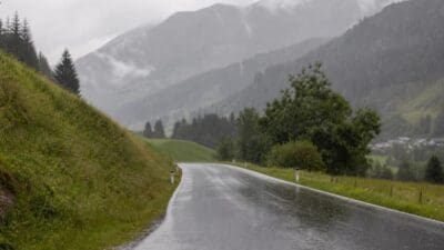 Nasse Landstraße im Regen vor bewaldeten Bergen und grünen Hängen