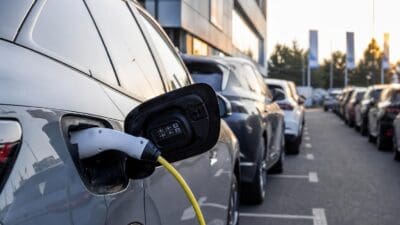 Silbernes Elektroauto mit offenem Ladeanschluss und eingestecktem Kabel an einer Ladestation, dahinter parkende Autos vor modernem Gebäude