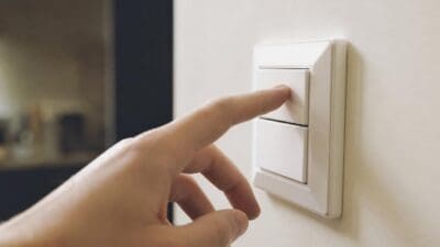 Eine Hand betätigt den oberen Schalter eines weißen Lichtschalters an der Wand.