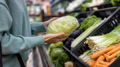 Eine Person hält einen in Plastikfolie verpackten Salatkopf in der Gemüseabteilung eines Supermarkts.