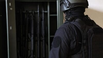 Einsatzkräfte in schwarzer Schutzausrüstung mit Helm vor offenem Waffenschrank mit Gewehren